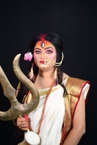 A person dressed in traditional attire resembling a deity figure, holding a trident in one hand and wearing colorful makeup and jewelry. The costume includes a white saree with gold and red borders, a headpiece with a third eye design, and a flower in the hair.