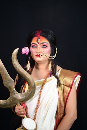 A person dressed in traditional attire resembling a deity figure, holding a trident in one hand and wearing colorful makeup and jewelry. The costume includes a white saree with gold and red borders, a headpiece with a third eye design, and a flower in the hair.
