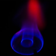 A vivid display of a gas stove burner with a glowing blue flame surrounded by a dark background. The flame is primarily blue with hints of purple, and it appears to transition to red toward the top, creating a warm and striking contrast.