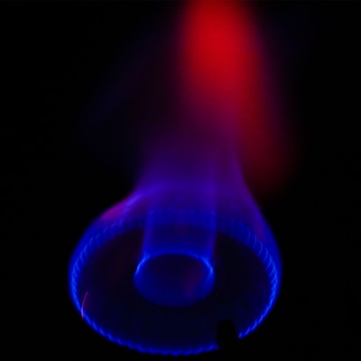 A vivid display of a gas stove burner with a glowing blue flame surrounded by a dark background. The flame is primarily blue with hints of purple, and it appears to transition to red toward the top, creating a warm and striking contrast.