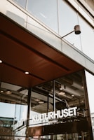 A modern architectural structure featuring large glass windows and a prominent entrance. The signage reads 'KULTURHUSET' in bold white letters. Industrial details like exposed beams and ceiling lights are visible through the windows, conveying an urban and contemporary atmosphere.
