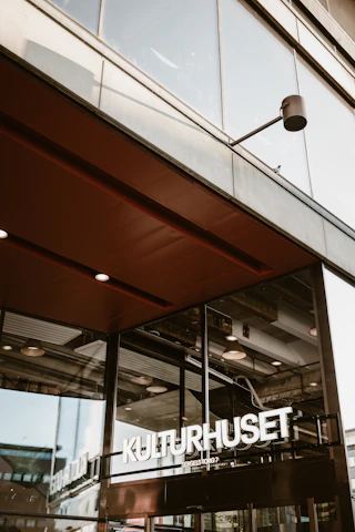 A modern architectural structure featuring large glass windows and a prominent entrance. The signage reads 'KULTURHUSET' in bold white letters. Industrial details like exposed beams and ceiling lights are visible through the windows, conveying an urban and contemporary atmosphere.