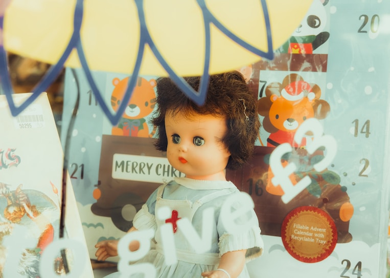 A vintage doll with curly brown hair and blue eyes dressed in a white outfit stands in front of a glass display. Behind it is an advent calendar with cartoon animals and Christmas decorations. There are hand-painted signs on the glass reading 'give £3'. The background includes playful imagery such as a tiger and bear with Christmas hats.