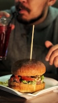 A close-up of a large hamburger placed on a square white plate, filled with lettuce, tomato, and a beef patty, with a wooden skewer piercing through it. In the background, a person in a gray sweater sips a red drink through a straw.