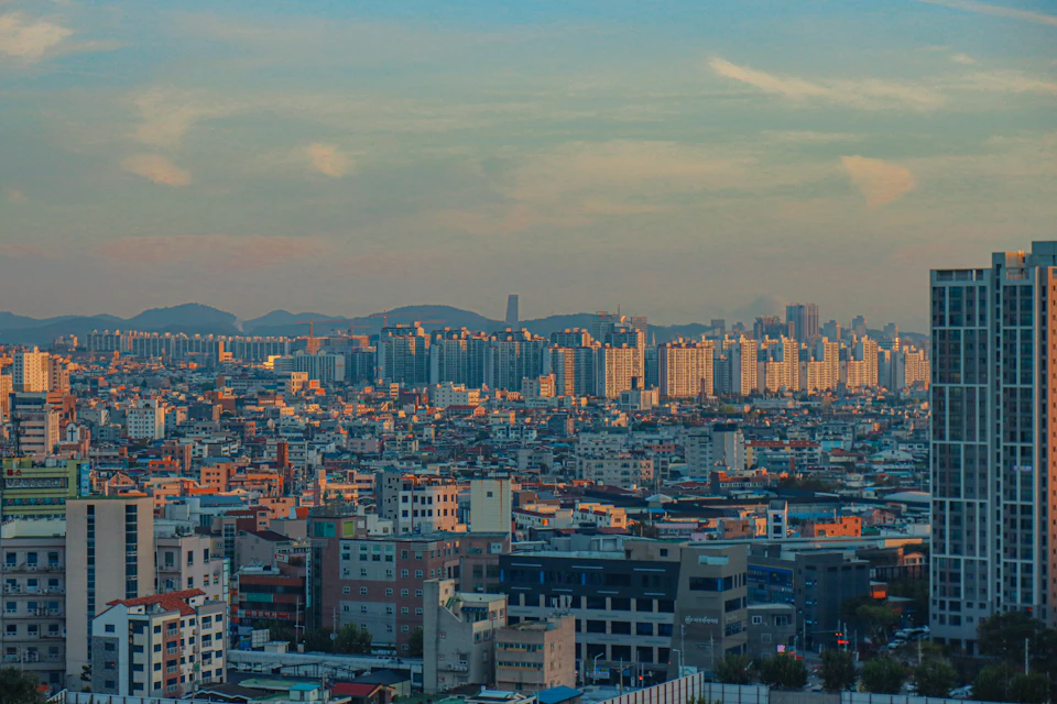 The image presents a panoramic view of a densely populated cityscape, with numerous tall buildings and skyscrapers reaching towards the sky. The city is bathed in warm hues from the setting sun, casting long shadows and bathing the urban landscape in a golden glow. The perspective of the photo is taken from a high vantage point, providing an expansive view of the city's skyline against the backdrop of the clear blue sky dotted with wispy clouds.

The buildings vary in height and architectural style, showcasing the diversity and vibrancy of the city. Some structures are taller than others, creating a sense of depth and scale within the image. The sun is positioned low on the horizon, casting long shadows that add to the dramatic effect of the scene.