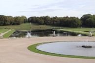 A serene landscape featuring a large, formal garden with circular and rectangular water features, surrounded by manicured lawns and dense greenery. Statues and fountains adorn the paths beside the water, with trees lining the background under a partly cloudy sky.