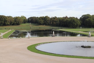A serene landscape featuring a large, formal garden with circular and rectangular water features, surrounded by manicured lawns and dense greenery. Statues and fountains adorn the paths beside the water, with trees lining the background under a partly cloudy sky.