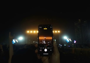 A hand is holding a smartphone capturing a live concert performance. The stage is lit with bright yellow lights, and the screen of the phone shows the stage slightly zoomed in. The audience is visible as silhouettes against the stage lighting, and another hand in the background appears to be making a rock hand sign.