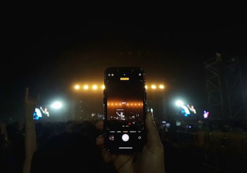 A hand is holding a smartphone capturing a live concert performance. The stage is lit with bright yellow lights, and the screen of the phone shows the stage slightly zoomed in. The audience is visible as silhouettes against the stage lighting, and another hand in the background appears to be making a rock hand sign.