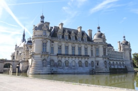 A grand, historical chateau with ornate architectural details and numerous large windows. The chateau is surrounded by a moat, reflecting its stone structure in the water. The sky is clear with visible contrails.