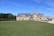 A grand chateau with intricate architecture, featuring multiple spires and detailed stonework, is surrounded by a vast expanse of lush green lawn. Trees line the background alongside a small body of water, creating a serene and peaceful setting under a clear blue sky.
