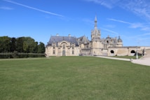 A grand chateau with intricate architecture, featuring multiple spires and detailed stonework, is surrounded by a vast expanse of lush green lawn. Trees line the background alongside a small body of water, creating a serene and peaceful setting under a clear blue sky.