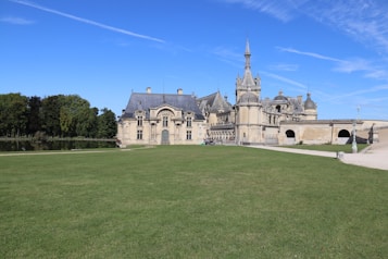 A grand chateau with intricate architecture, featuring multiple spires and detailed stonework, is surrounded by a vast expanse of lush green lawn. Trees line the background alongside a small body of water, creating a serene and peaceful setting under a clear blue sky.