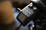 A close-up view of an audio recording device with multiple buttons and a screen. The device is mounted on a tripod, and a part of the tripod and its some of its components are visible. There is also a piece of black equipment or strap with the word 'STUDIO' partially visible in the upper right corner.