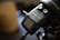 A close-up view of an audio recording device with multiple buttons and a screen. The device is mounted on a tripod, and a part of the tripod and its some of its components are visible. There is also a piece of black equipment or strap with the word 'STUDIO' partially visible in the upper right corner.