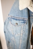 A close-up view of a light blue denim jacket featuring prominent stitching details, metal button closures, and a white faux fur collar. The texture of the denim fabric is clearly visible, providing a casual and comfortable appearance.