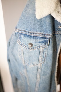 A close-up view of a light blue denim jacket featuring prominent stitching details, metal button closures, and a white faux fur collar. The texture of the denim fabric is clearly visible, providing a casual and comfortable appearance.