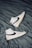 A pair of white high-top sneakers with black swoosh logos are placed on a crumpled dark gray fabric surface. The sneakers are positioned in opposite directions, with one shoe pointing upwards and the other downwards. The overall design is sleek and modern, with subtle beige accents on the soles.