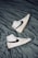 A pair of white high-top sneakers with black swoosh logos are placed on a crumpled dark gray fabric surface. The sneakers are positioned in opposite directions, with one shoe pointing upwards and the other downwards. The overall design is sleek and modern, with subtle beige accents on the soles.