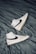 A pair of white high-top sneakers with black swoosh logos are placed on a crumpled dark gray fabric surface. The sneakers are positioned in opposite directions, with one shoe pointing upwards and the other downwards. The overall design is sleek and modern, with subtle beige accents on the soles.