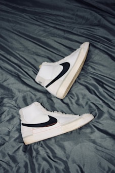 A pair of white high-top sneakers with black swoosh logos are placed on a crumpled dark gray fabric surface. The sneakers are positioned in opposite directions, with one shoe pointing upwards and the other downwards. The overall design is sleek and modern, with subtle beige accents on the soles.