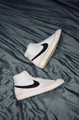 A pair of white high-top sneakers with black swoosh logos are placed on a crumpled dark gray fabric surface. The sneakers are positioned in opposite directions, with one shoe pointing upwards and the other downwards. The overall design is sleek and modern, with subtle beige accents on the soles.