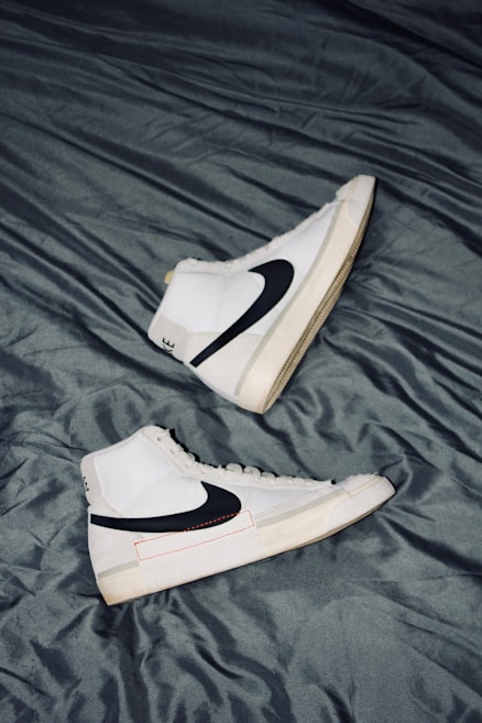 A pair of white high-top sneakers with black swoosh logos are placed on a crumpled dark gray fabric surface. The sneakers are positioned in opposite directions, with one shoe pointing upwards and the other downwards. The overall design is sleek and modern, with subtle beige accents on the soles.