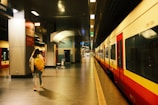 A person with a yellow backpack walks along a train platform inside a modern station. The platform is empty, with bright overhead lighting illuminating the area. A train, primarily red and white in color, is stationed to the right. Signage on the walls indicates directions, and a luggage cart is visible in the background.