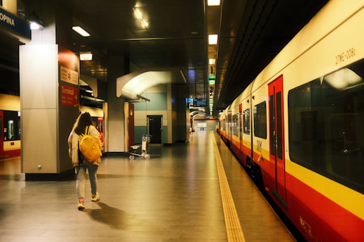 A person with a yellow backpack walks along a train platform inside a modern station. The platform is empty, with bright overhead lighting illuminating the area. A train, primarily red and white in color, is stationed to the right. Signage on the walls indicates directions, and a luggage cart is visible in the background.