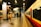 A person with a yellow backpack walks along a train platform inside a modern station. The platform is empty, with bright overhead lighting illuminating the area. A train, primarily red and white in color, is stationed to the right. Signage on the walls indicates directions, and a luggage cart is visible in the background.
