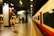 A person with a yellow backpack walks along a train platform inside a modern station. The platform is empty, with bright overhead lighting illuminating the area. A train, primarily red and white in color, is stationed to the right. Signage on the walls indicates directions, and a luggage cart is visible in the background.