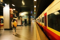 A person with a yellow backpack walks along a train platform inside a modern station. The platform is empty, with bright overhead lighting illuminating the area. A train, primarily red and white in color, is stationed to the right. Signage on the walls indicates directions, and a luggage cart is visible in the background.