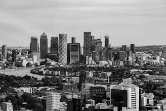 A black and white photograph of a modern city skyline with tall skyscrapers and a river in the foreground. The skyline features distinctive high-rises and a dense cityscape. The image is captured from an elevated perspective, showing urban architecture and sprawling buildings.