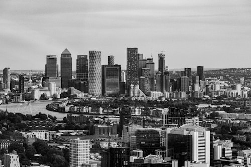 A black and white photograph of a modern city skyline with tall skyscrapers and a river in the foreground. The skyline features distinctive high-rises and a dense cityscape. The image is captured from an elevated perspective, showing urban architecture and sprawling buildings.