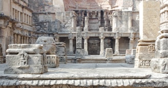 An intricately carved stone structure featuring numerous columns and detailed relief work on the walls. The architecture suggests an ancient or historical site with weathered stones and a mix of angular and rounded shapes. The scene appears to be part of a larger complex, showcasing classical architecture elements with artistic designs etched into the stone.