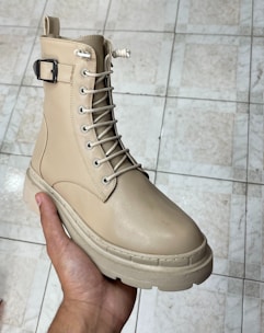 A hand is holding a beige boot with a buckle and lace-up design, showcasing a rugged sole. The footwear appears well-crafted and is placed against a tiled floor, likely indoors.