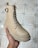 A hand is holding a beige boot with a buckle and lace-up design, showcasing a rugged sole. The footwear appears well-crafted and is placed against a tiled floor, likely indoors.