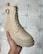 A hand is holding a beige boot with a buckle and lace-up design, showcasing a rugged sole. The footwear appears well-crafted and is placed against a tiled floor, likely indoors.