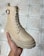 A hand is holding a beige boot with a buckle and lace-up design, showcasing a rugged sole. The footwear appears well-crafted and is placed against a tiled floor, likely indoors.