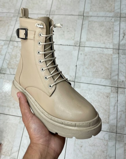 A hand is holding a beige boot with a buckle and lace-up design, showcasing a rugged sole. The footwear appears well-crafted and is placed against a tiled floor, likely indoors.