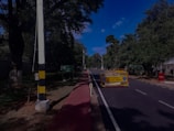 A road lined with trees on either side is partially blocked by a barrier indicating it is an accident-prone area. The sky is clear and blue, and there is a sidewalk alongside the road.