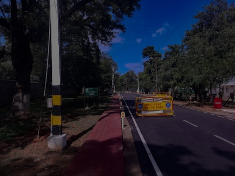 A road lined with trees on either side is partially blocked by a barrier indicating it is an accident-prone area. The sky is clear and blue, and there is a sidewalk alongside the road.