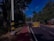 A road lined with trees on either side is partially blocked by a barrier indicating it is an accident-prone area. The sky is clear and blue, and there is a sidewalk alongside the road.