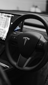 A sleek, modern steering wheel featuring the Tesla logo. The interior of the vehicle includes a large touchscreen display showing a map interface, suggesting advanced technology and navigation capabilities.