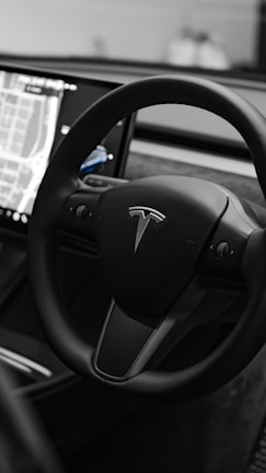 A sleek, modern steering wheel featuring the Tesla logo. The interior of the vehicle includes a large touchscreen display showing a map interface, suggesting advanced technology and navigation capabilities.