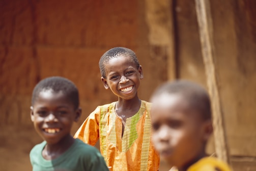 Three children are smiling and looking at the camera. The child in the center is wearing an orange traditional outfit with yellow stripes and standing out sharply, while the other two children are slightly blurred in the foreground. The background appears to be an outdoor setting with earthy tones and a wooden structure.