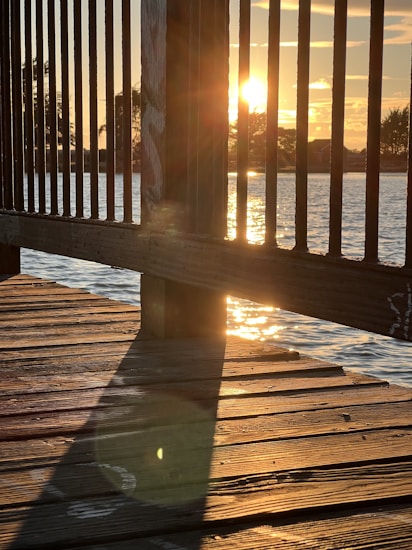 The image captures a sunset viewed from a wooden dock. The sun is partially visible through the vertical slats of a railing, casting a warm golden hue over the scene. Reflections of the sun can be seen on the water surface, and shadows of the railing are cast on the weathered wooden planks of the dock. Trees and a house are faintly visible in the background.