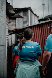 A group of people is seen from behind, wearing blue shirts with 'LA VIDA' printed on them, walking through an alley lined with corrugated metal structures. The atmosphere appears overcast and subdued.