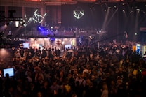 A large crowd of people gathered at a bustling event or conference with various booths and lighting installations. The atmosphere is lively and busy, with attendees engaging in discussions and exploring the exhibits. Overhead lighting and geometric designs add a modern, tech-inspired ambiance.