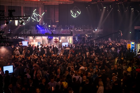 A large crowd of people gathered at a bustling event or conference with various booths and lighting installations. The atmosphere is lively and busy, with attendees engaging in discussions and exploring the exhibits. Overhead lighting and geometric designs add a modern, tech-inspired ambiance.